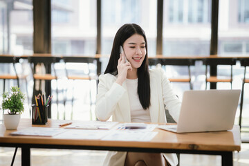 Beautiful Asian female accountant working with smartphone and laptop at her desk analyzing business reports and documents. Sending messages