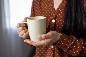 Close-up of asian woman holding mug, displaying comfort, warmth, and relaxation. Hands cradling neutral-colored cup, wearing red polka dot blouse, suggesting cozy, calm atmosphere.