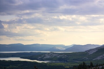 A scenic landscape view of a lake surrounded by mountains and forests under a cloudy sky