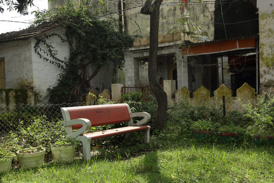A quiet, charming spot with a red bench amidst lush greenery, set beside an old, vine-covered house. The scene exudes a sense of calm and nature&rsquo;s embrace.