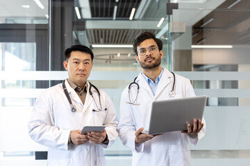 Doctors in white coats collaborating in modern office, using tablet and laptop for healthcare discussions. Stethoscopes visible, highlighting medical professionals' teamwork and technology.