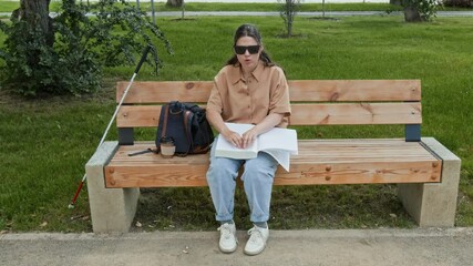 Blind young woman with dark glasses reading braille book while resting on bench in park on summer day