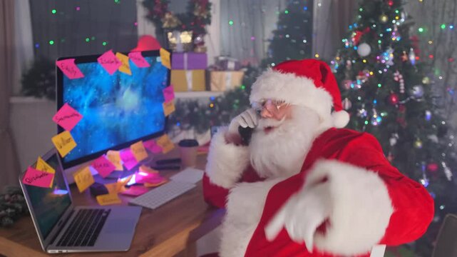Santa actively gestures on the phone while multitasking on his computer, fully immersed in the busy Christmas preparations. 