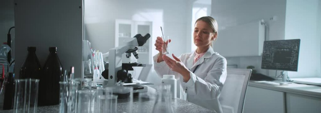 In the laboratory scientist woman looking under the microscope holds a tube at sunlight. Medicine, biotechnology research. Pharmacology
