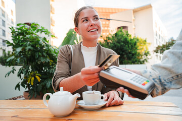 Smiling young woman sitting at a bar terrace, paying for her tea using a credit card and contactless payment terminal, highlighting convenience and modern technology in outdoor dining settings