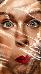 Close-up of woman face with wide eyes expressing surprise while her face being wrapped in transparent plastic. Grimacing. Conceptual photography. Concept of human emotions