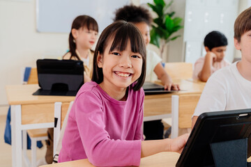 Young Asian girl smiling and looking at camera with tablet in diverse elementary classroom. Students embrace digital learning, showcasing modern education techniques and technology