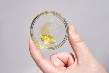 Hand holding clear glass cup with yellow liquid capsule on light gray background for supplement or health concept.