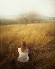 a girl in a white dress, sitting in a field on yellow grass. She has her back to the audience