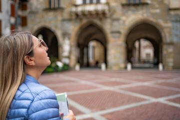 Piazza Vecchia, Bergamo Italy. Beautiful young woman wearing blue jacket and sunglasses admiring main square in old town, holding map in her hand and looking up, on autumn day. Side view. City tour.