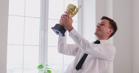 Happy businessman standing at office, holding business champion award. Joyful male leader celebrating great hard work success, shaking golden trophy cup and kissing it. Business goals achievement. - Powered by Adobe