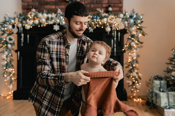 Fototapeta premium A young father helps his son change his sweater near Christmas tree in living room. Concept of solo parenting and gifts for New Year, Mother's Day and Father's Day. Modern interior. Part of a series