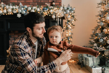 A young father helps his son change his sweater near Christmas tree in living room. Concept of solo parenting and gifts for New Year, Mother's Day and Father's Day. Modern interior. Part of a series