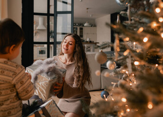 A happy single mother gives New Year's gifts to her son near Christmas tree in living room. Concept of solo parenting and gifts for New Year, Mother's Day and Father's Day. Part of a series