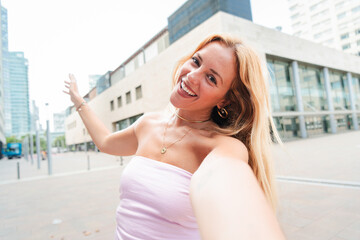Blonde Caucasian woman taking a cheerful selfie while flashing a white smile in a modern urban...