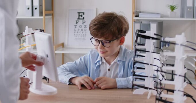 Ophthalmologist doctor helps little boy choose glasses for vision in eye clinic. Smiling boy sits at table among various types of glasses, tries on different frames and looks in mirror. Video footage.