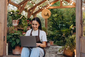 Happy asian lady sitting with portable computer by doors of greenhouse and surfing information...