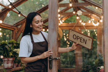 Fototapeta premium Optimistic brunette with smiling face setting sign open hanging on glassy door of botany store