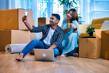 Indian Asian couple clicking selfie while sitting between moving boxes during relocation