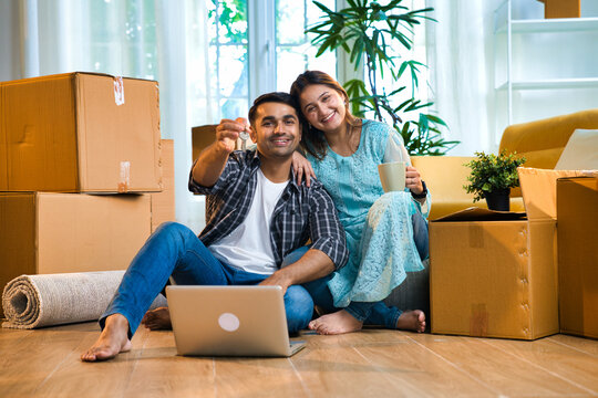 Young Indian Asian couple sitting with moving boxes, joyfully showing keys to their new apartment