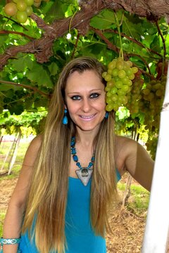 Mulher loira, cabelos lisos e longos, olhos azuis, no meio de um parreiral de uvas