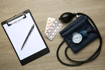 Blood pressure measuring device, pills and clipboard on wooden table, top view
