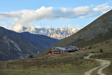 Fototapeta premium Picturesque mountain landscape. A narrow gorge with a rocky mountain under a white cloud in the distance. A chalet with a winding road to it in the foreground.