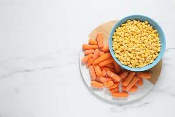 Frozen corn kernels in bowl and baby carrots on white marble table, top view. Space for text