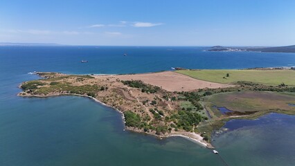 A stunning aerial view of a coastal landscape featuring a serene bay, vast green fields, and distant mountains under a clear blue sky. The image captures a peaceful seaside area with a few ships