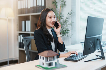 Young real estate agent talking on the phone and working on a computer with a house model on the table