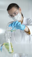 Woman scientist, wearing a lab coat, blue gloves and protective glasses, is pouring a yellow liquid from the beaker into Erlenmeyer flask in laboratory, portrait and vertical view. Medicine, science