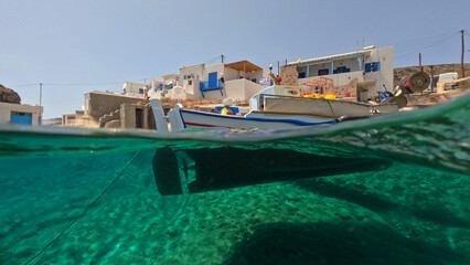 Underwater split photo of traditional fishing boat anchored in small cove of Goupa Kara famous for colourful boat houses called Sirmata, Kimolos island, Cyclades, Greece