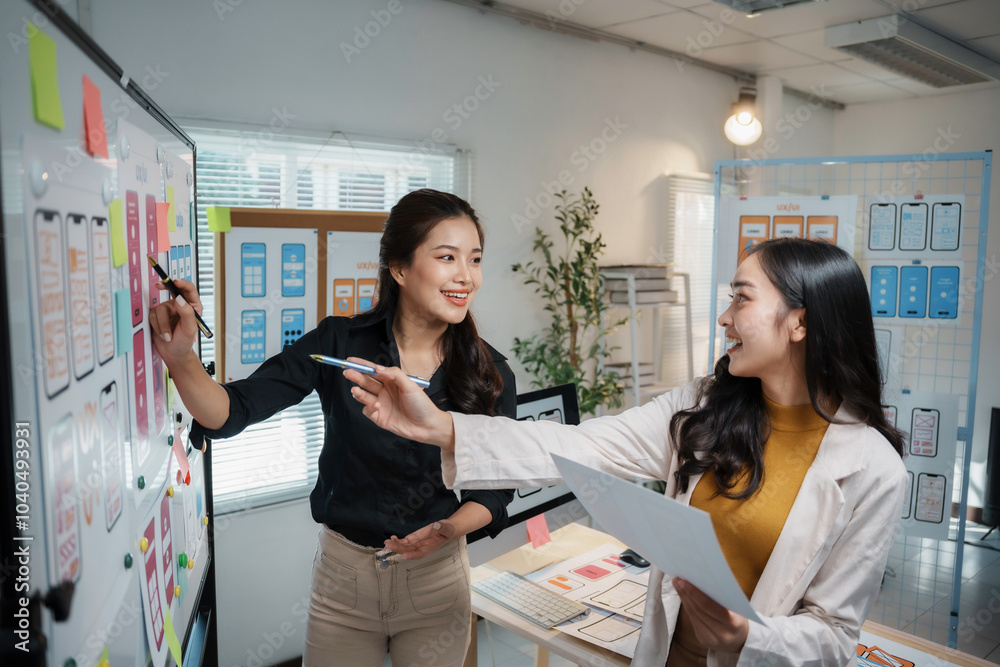 Wall mural two young asian uiux designers are happily discussing mobile app wireframe design displayed on a whi