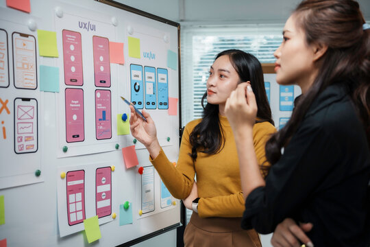 Two asian uiux designers discussing over mobile app interface wireframe design with laptop computer at modern office