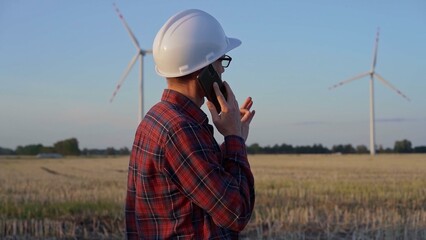 Man engineer, wearing a white protective helmet is talking by smartphone in a field with wind turbines, as the sun sets. Clean energy and engineering concept