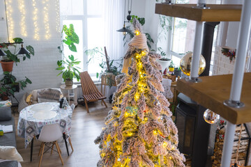 Festive Christmas decor in white kitchen, modern village interior with a snowy Christmas tree and fairy lights. New Year, Christmas mood, cozy home, Metal modular staircase. The general plan