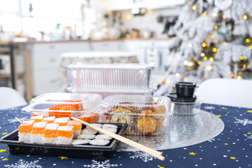 Delivery of sushi and rolls in food containers on the table in the festive interior of the kitchen of the house with a white Christmas tree for Christmas and New year. Food delivery for the holidays