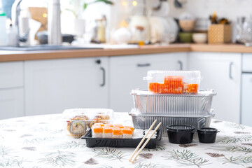 Delivery of sushi and rolls in food containers on the table in the festive interior of the kitchen of the house with a white Christmas tree for Christmas and New year. Food delivery for the holidays