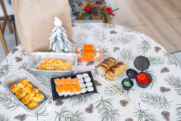 Delivery of sushi and rolls in food containers on the table in the festive interior of the kitchen of the house with a white Christmas tree for Christmas and New year. Food delivery for the holidays