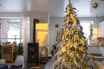 Festive white modern interior of house is decorated for Christmas and New Year in loft style with black stove, fireplace, Christmas tree. Potted plants, firewood in the woodpile