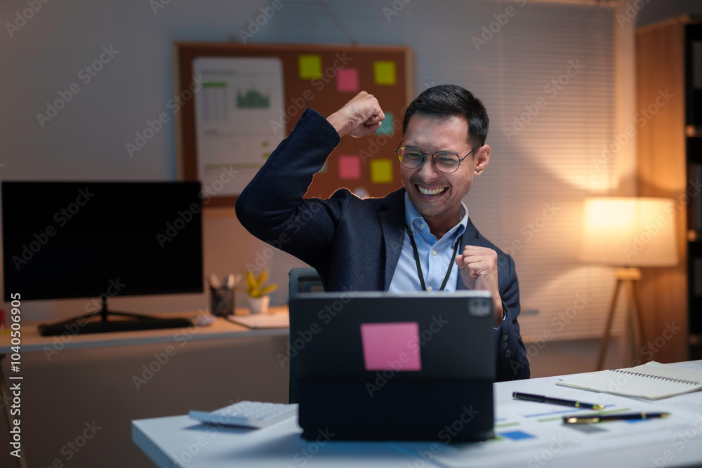 Wall mural asian businessman is raising his arms in the air while sitting at his desk working late in the offic