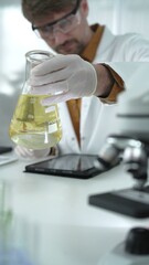 Man scientist, wearing white gloves and protective glasses, is holding a yellow chemical solution...
