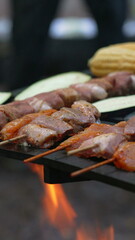 Close-up of marinated meat skewers grilling on a campfire with corn. The scene focuses on the texture and details of the food, evoking a sense of outdoor culinary experience