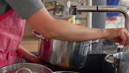 Hands filling a metal pot with water from the kitchen faucet. The scene focuses on the water stream...