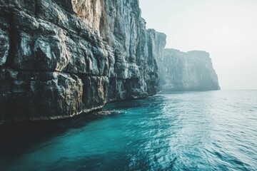 Misty cliffs rise majestically over the deep blue ocean under an overcast sky, offering a moody and...