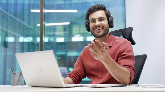 Smiling IT specialist in wireless headphones asks to join him, beckons with inviting hand gesture while sitting at laptop at workplace in office. Friendly developer or coder invites to join the team