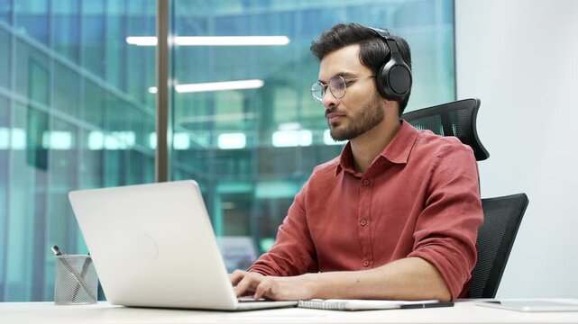 Portrait of a happy handsome IT specialist in wireless headphones working on a laptop sitting at a workplace in a business office. Confident successful smiling developer or coder looking at camera