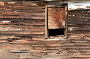 Old wooden exterior wall of a building with window partly boarded up shows weathered siding with no paint showing.