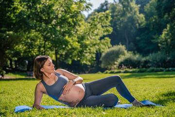 Pregnant woman exercising in park enjoying warm summer sun practicing yoga on green grass