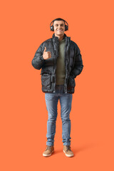 Young man in winter clothes with headphones showing thumb-up on orange background